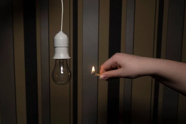 A hand with a burning match next to a holder with an electric light bulb in a dark room (close-up). Blackout. Power outage concept