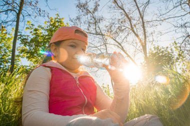 Bir kız, kalın yeşil çimenlerde oturur ve gün batımının arka planına karşı şişeden su içer. Şehir parkında dışarı çık.