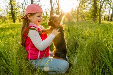 Neşeli bir kız köpeğiyle birlikte kalın yeşil çimlerde oturur gün batımının arka planına karşı. Şehir parkında dışarı çık.