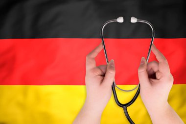 Alman bayrağının arka planına karşı tıbbi steteskop elinde (yakın plan). Alman sağlık sistemi kavramı