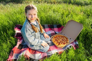 Mavi kot takım elbiseli bir kız elinde bir kutu pizzayla yeşil çimlerin üzerinde bir battaniyenin üzerinde oturuyor. Yaz güneşli bir gün. Açık hava pikniği