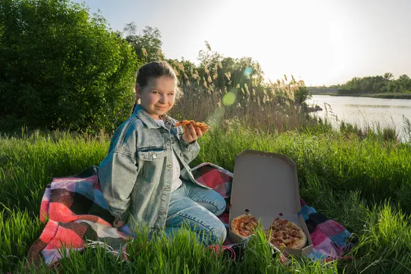 A girl in a blue denim suit sits on a blanket on green grass on the river bank and holds a slice of pizza in her hand. Summer sunny day. Outdoor picnic