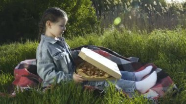 Mavi kot takım elbiseli bir kız yeşil çimlerin üzerinde bir battaniyeye oturur ve bir kutu pizza açar. Yaz güneşli bir gün. Açık hava pikniği