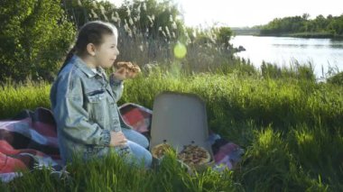 Mavi kot takım elbiseli bir kız yeşil çimlerin üzerinde bir battaniyeye oturur ve bir dilim pizza yer. Yaz güneşli bir gün. Açık hava pikniği