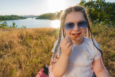 Mavi güneş gözlüklü bir kız nehir kıyısında oturur ve patates kızartması yer. Kırsalda güneşli bir yaz günü. Açık hava pikniği