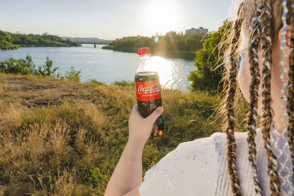 Krivoy Rog, Ukrayna, 26 Haziran 2024: Bir kız nehrin kıyısında duruyor ve elinde bir şişe Coca-Cola tutuyor (arkadan görüş). Yaz güneşli bir gün. Açık hava eğlencesi
