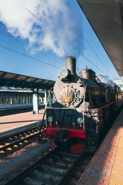Mavi gökyüzünün altındaki istasyon platformunda duran tarihi buharlı tren lokomotifi. Buhar yaymak, klasik ulaşım ve tarihi temsil etmek