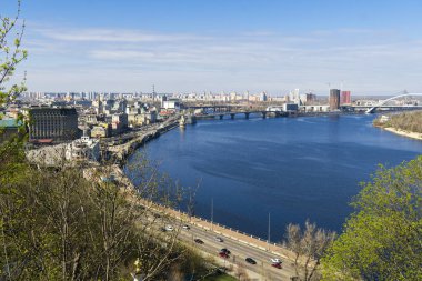Kyiv 'in panoramik yüksek açılı görüntüsü geniş bir Dnipro nehrinin şehrin içinden geçişi, birden fazla köprü ve mavi gökyüzü altında tarihi ve modern mimarinin bir karışımı.
