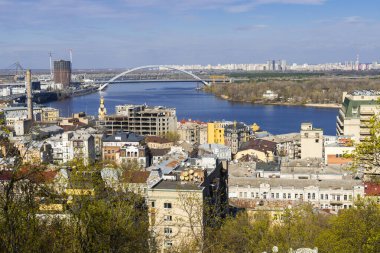 Kyiv 'in şehir manzarasının panoramik bir görüntüsü, eski ve yeni binaların karışımı, geniş bir Dnipro nehri, ve parlak mavi gökyüzünün altında belirgin bir kemer köprüsü.