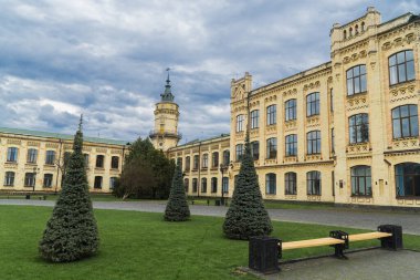 Geniş, sarı tuğla bina, detaylı mimari özelliklere sahip, sayısız pencere, ve bulutlu bir günde çevreleyen yeşil çimenler ve ağaçlar.