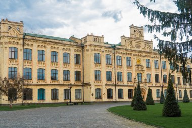 Geniş, sarı tuğla bina, detaylı mimari özelliklere sahip, sayısız pencere, ve bulutlu bir günde çevreleyen yeşil çimenler ve ağaçlar.