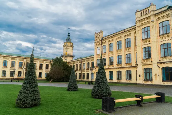 Geniş, sarı tuğla bina, detaylı mimari özelliklere sahip, sayısız pencere, ve bulutlu bir günde çevreleyen yeşil çimenler ve ağaçlar.