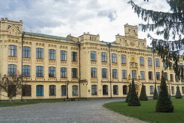 Geniş, sarı tuğla bina, detaylı mimari özelliklere sahip, sayısız pencere, ve bulutlu bir günde çevreleyen yeşil çimenler ve ağaçlar.