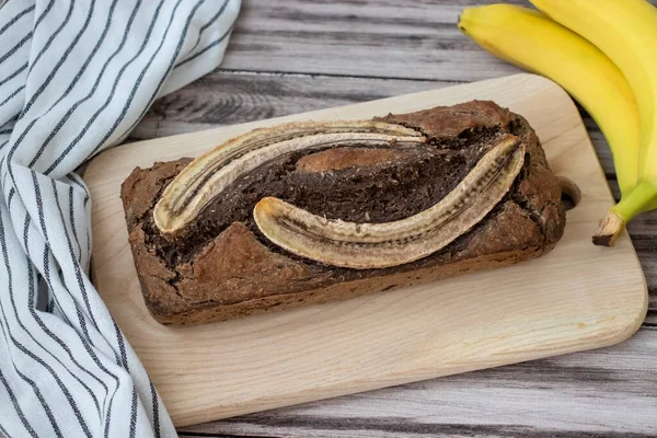 Brown banana bread with banana halves on top on wooden surface with fresh bananas.