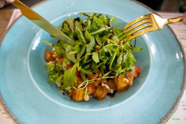 Arugula salatası, karamelli patlıcan ve mavi tabakta çatal bıçak..