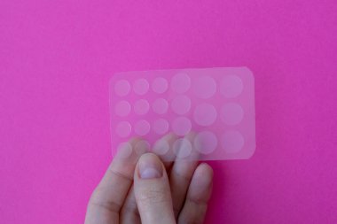 Pembe arka planda sivilce yamaları. Bir kadının elinde şeffaf yüz bantları var.