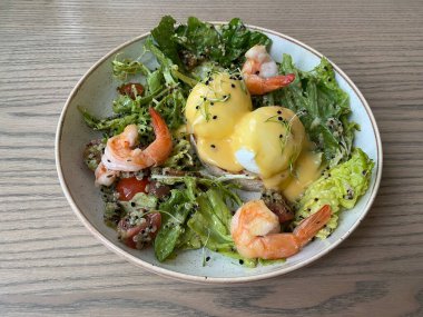 Sağlıklı LCHF kahvaltısı. Kinoalı Benedict yumurtası, taze salata, karides ve sos ahşap bir masada bej bir tabakta.