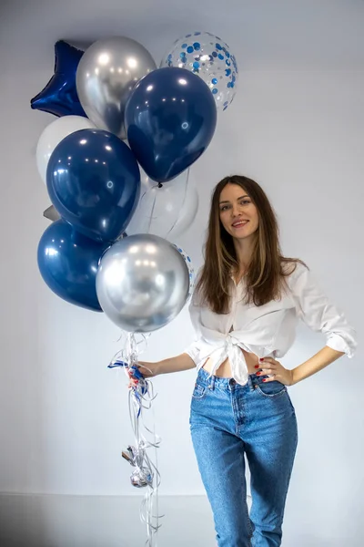 Party Time. A charming woman in a stylish shirt and jeans is holding a bunch of blue balloons against a white background. A joyful young woman is having fun at the party, enjoying the festivities.