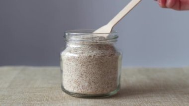 Psilyum bir muz kabuğudur. Cam bir kavanoz içinde ahşap bir kaşık olan Psyllium 'un bej ve gri arka planda serpiştirdiği