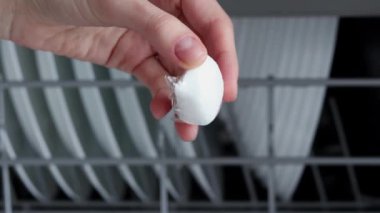 Bulaşıkçı tableti, açık bir bulaşık makinesinin önünde, elinde temiz bulaşıklar olan bir kadının elinde. Beyaz kapsül suda çözünebilir ambalajda. Ev için eko ürünler.
