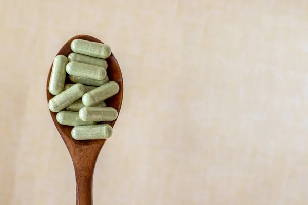 Transparent green powder tablet capsules in a wooden spoon on a beige background. Dietary fiber prebiotic supplements for healthy gut. Copy space