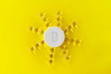 Sarı arka planda sarı ilaç kapsülü hapları ve sarı arka planda güneş şeklinde dizilmiş D vitamini içeren vatozlar var...