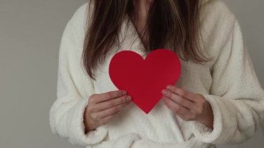 A woman in a robe holds a red paper heart. The concept of love or donation.