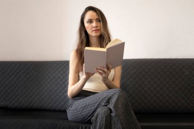 Beautiful, brooding young woman sits on the couch, holds an open book, and looks away. clipart