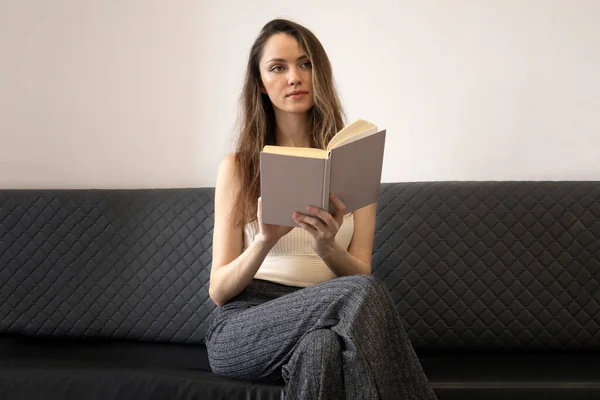 Stock image Beautiful, brooding young woman sits on the couch, holds an open book, and looks away.