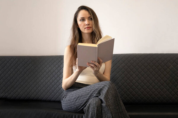 Beautiful, brooding young woman sits on the couch, holds an open book, and looks away.