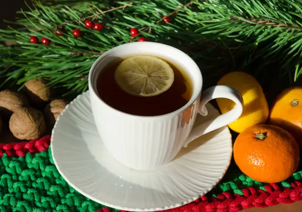 A cup of morning tea with lemon on a background of nuts, tangerines and green branches of a Christmas tree. Decorations for the New Year and Christmas. A place for congratulations.