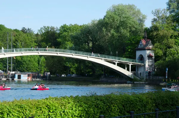Kendine özgü kulesi olan bir yaya köprüsü, Berlin 'in Treptower Parkı' ndaki göletin üzerinde zarifçe kıvrılır, açık mavi gökyüzünün altında yemyeşil ağaçlarla çevrili birçok pedallı tekne görülebilir..