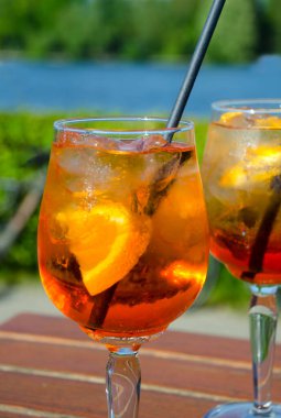 İki bardak parlak turuncu Aperol Spritz, yoğunlaşmayla kaplı, parlak portakal dilimi ve buz ile süslenmiş. Caddedeki bir kafede, bir nehrin ve yeşilliğin arka planında..