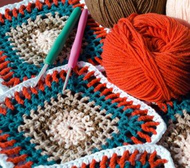 Örgü örülmüş bir nesnenin canlı bir yakın çekimi ve onun bitmiş ürünü. Parlak turuncu, turkuaz, kahverengi ve açık pembe iplik topları bir yığın rengarenk büyükanne karesinin yanına saçılmış..