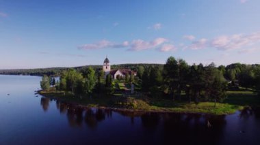 Aerial establishing shot over Lake Siljan, pull back shot of a 15th century church and cemetery. Historical monument on the lakeside. Mountains and forests in the background, during sunrise golden hour. In Rattvik, Dalarna Sweden