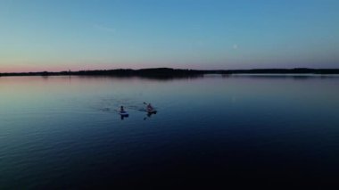 Young women enjoy on a SUP board during an leisure activity at a lake. Blue hour sunset sky in sweden. Healthy lifestyle. Water sports, SUP surfers adventure summer holiday. Low angle panning shot