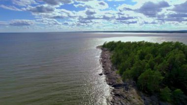 Aerial view above a remote rocky islet in the middle of a lake with small waves, blue sky with white fluffy clouds. Mountains and forests on the horizon. Drone static aerial view, Saffle, Vanern lake, Varmland, Sweden