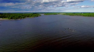 Low aerial backwards over rocky island with pine trees. Islet in the middle of the lake with water waves, cloudy blue sky. Vanern lake in Saffle Varmland Sweden