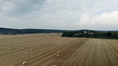 Hasattan sonra saman balyalarıyla çevrili altın sarısı tarlalarla çevrili bir kilisenin yay görüntüsü. Bulutlu bir gökyüzünde arka plandaki orman ve tepelerde. Gammalstorp Kyrka İsveç. İHA yüksek açı görünümü