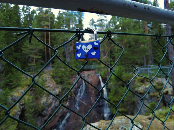 Blue padlock with hearts decoration and initials attached to fence at waterfall. Couple in love confirm with padlock eternal love symbol. In the background a waterfall and forest, shallow depth of field
