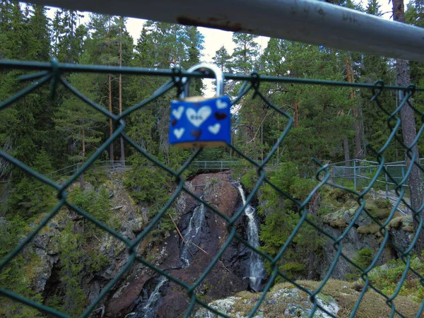 Blue padlock with hearts decoration attached to fence, out of focus. In the background a waterfall and forest in focus. Love confirm with padlock eternal love concept.