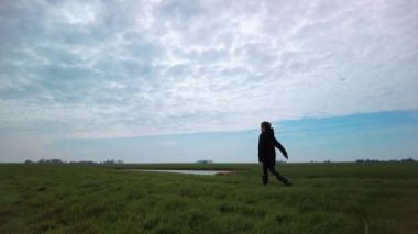 Child in winter coat and boots walks briskly steadfast alone over the green grass trail through the rural landscape and farmlands in early spring on a cloudy day. Low angle left side view, medium shot, slow motion.