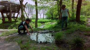 İki çocuk su birikintisinde çamur ve suyla oynuyor. Güneşli bir bahar günü parkta ağaçların gölgesinde oynayan çocuklar. Düşük açılı statik çekim, gerçek zamanlı.