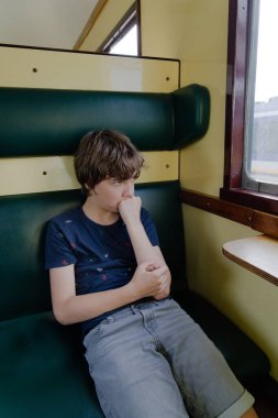 Çocuk yalnız oturuyor ve eski tren vagonunun camından dışarı bakıyor, çocuğun çenesine ve ağzına yumruk yemiş, ciddi bakışlar atan bir bakış açısı var.