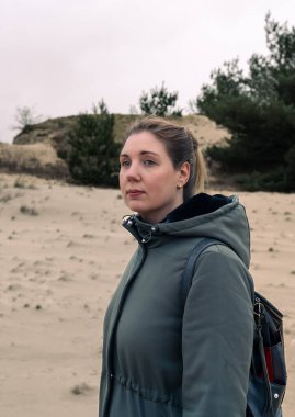 Bulutlu bir gökyüzünün altındaki kum tepelerinde dalgın genç bir kadının portresi. Sarışın, temiz bir şekilde geri çekilmiş. Düşünceli bir ifadeyle, yeşil, rahat bir ceketle uzaklardan bakıyor.