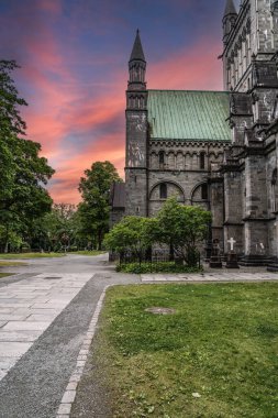 Norveç 'in Trondheim kentindeki Nidaros Katedrali avlusunda gün batımı. Akşam gökyüzü alevler içinde. Katedral kulesinin arkasında gün batımı renkleri var. Mezar taşları yemyeşil bahçeye saçılmış..