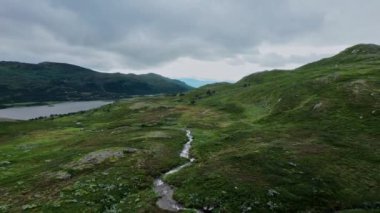 Bir yamacın üzerinde uçarken ve bir dağ deresi, yemyeşil bir Norveç dağı boyunca akar öğleden sonra bir gölün yanında, Norveç 'te açık bulutlu bir gökyüzü.