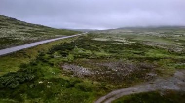 Sisli tundra dağ manzarasında bomboş bir yola paralel uçan insansız hava aracı Rondane Ulusal Parkı, Innlandet, Norveç 'te yeşil yosunlu bitki örtüsüyle kaplandı..