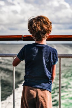 Bir gemi güvertesinde duran genç bir çocuğun arka görüntüsü, açık denizde gemi tarafından yaratılan kalıntılara bakıyor..