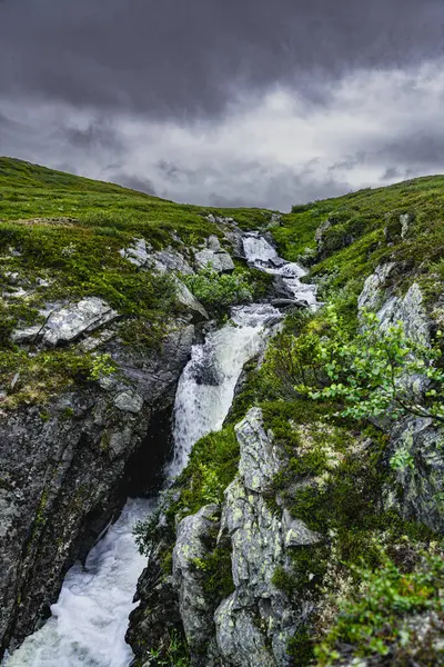 stock image A rocky mountainside in Norway features a stream of water flowing down, creating a cascading waterfall under a gray, cloudy sky